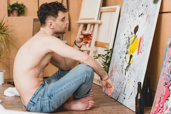 Bellissimo artista mezzo nudo seduto sul pavimento davanti all'immagine e con un bicchiere di whisky in mano — Foto stock