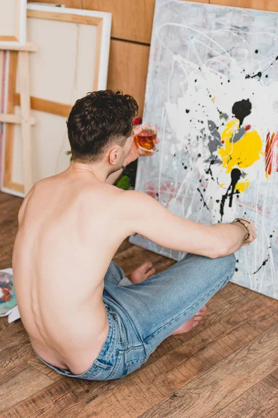 Half-naked artist sitting on floor in front of picture and holding glass of whiskey — Stock Photo