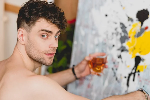 Selective focus of handsome artist holding glass of whiskey and looking at camera — Stock Photo