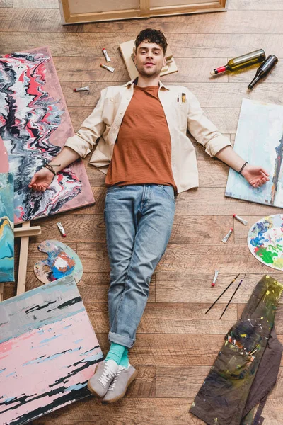 Tired artist lying of floor, surrounded with paintings and draw utensils, and looking at camera — Stock Photo