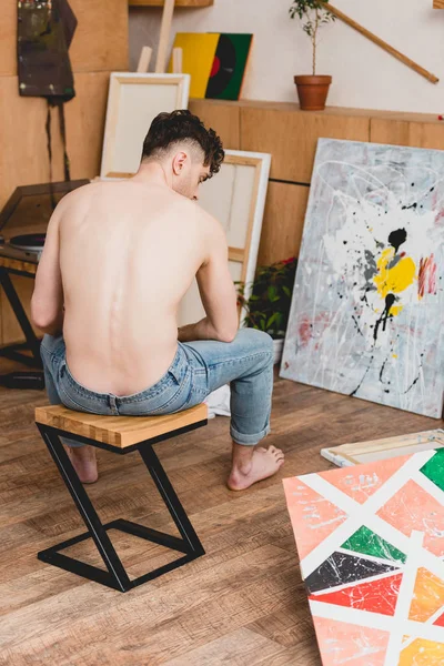 Полуголый художник в синих джинсах сидит на стуле в галерее, окруженный картинами — стоковое фото