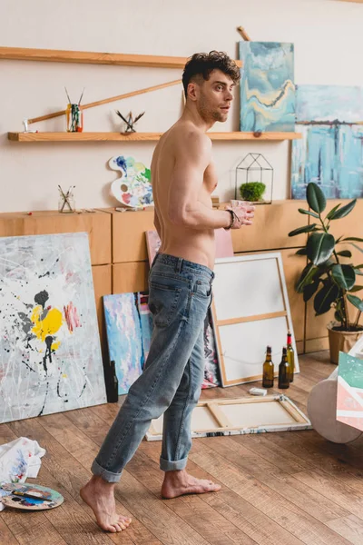 Beau artiste à moitié nu en jeans bleus debout dans l'atelier de peinture et regardant loin — Photo de stock