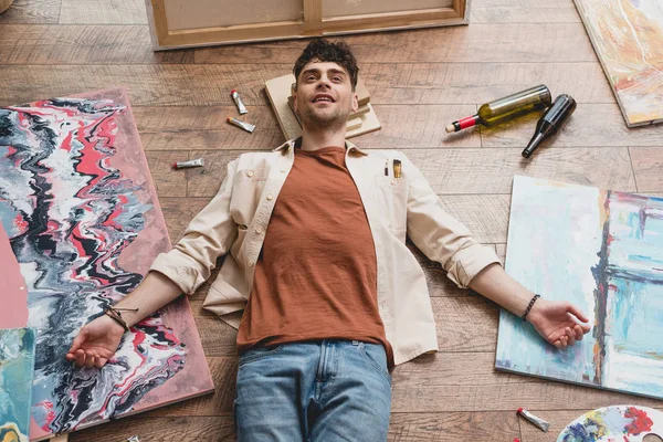 Artiste épuisé couché sur le sol dans un atelier de peinture, entouré de peintures, bouteilles vides et d'ustensiles de dessin — Photo de stock