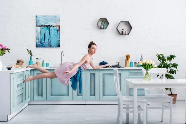 Bela menina descalça em vestido elegante e avental levitando no ar com panela na cozinha — Fotografia de Stock