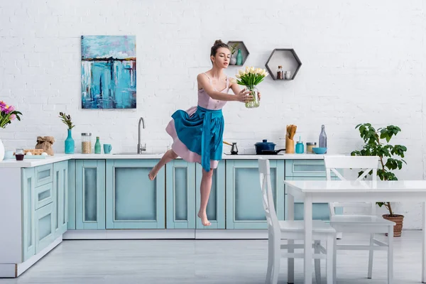 Hermosa chica descalza en vestido elegante y delantal volando en el aire con ramo de tulipanes en tarro de vidrio en la cocina - foto de stock