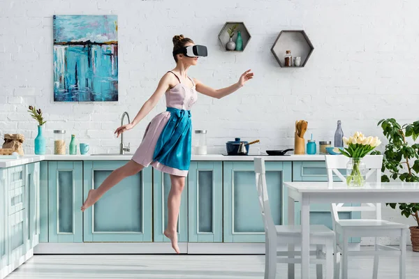 Молодая босиком женщина летит в воздухе в наушниках виртуальной реальности и жестом на кухне — стоковое фото