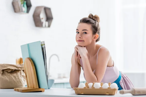 Glückliches attraktives Mädchen in der Nähe von Kochbuch und Eiern — Stockfoto