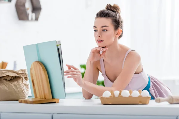 Attrayant fille lecture recette dans le livre de cuisine avec des ingrédients de boulangerie autour — Photo de stock