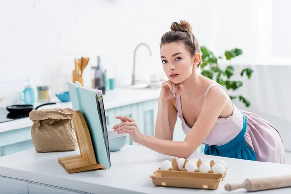 Chica atractiva tocando libro de cocina y mirando a la cámara con ingredientes de panadería alrededor - foto de stock