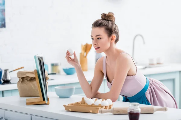 Menina atraente leitura receita no livro de receitas e segurando ovo na cozinha — Fotografia de Stock