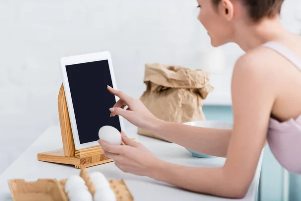 Abgeschnittene Ansicht einer jungen Frau, die ein digitales Tablet mit leerem Bildschirm benutzt, während sie ein Ei hält — Stockfoto