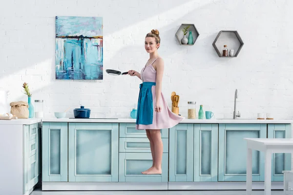 Hermosa mujer joven descalza en delantal levitando en la cocina mientras sostiene la cacerola - foto de stock