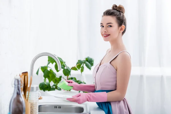 Feliz sorrindo jovem mulher em luvas de borracha lavar pratos na cozinha — Fotografia de Stock