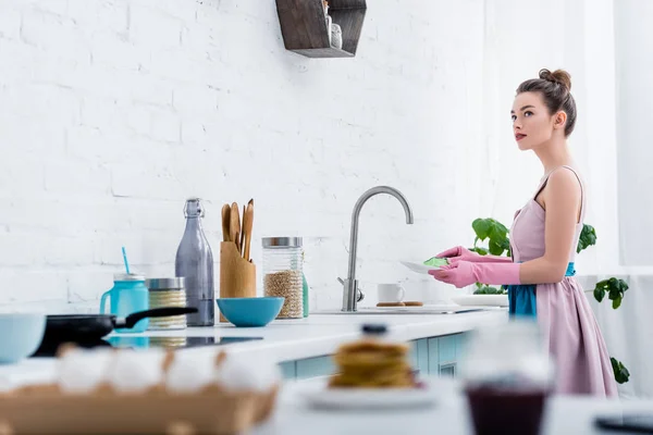 Messa a fuoco selettiva di giovane donna in guanti di gomma lavare i piatti in cucina — Foto stock