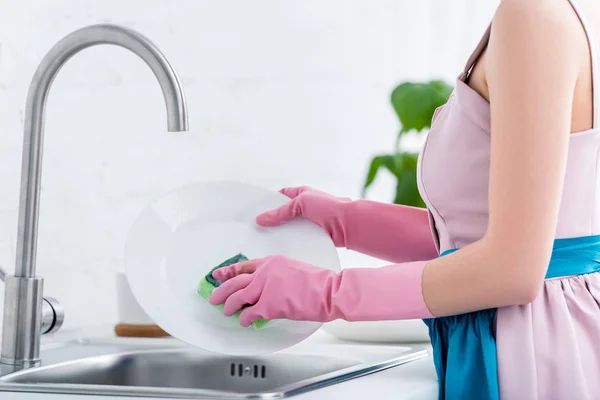 Vista recortada de mujer joven en guantes de goma rosa lavar los platos en la cocina - foto de stock
