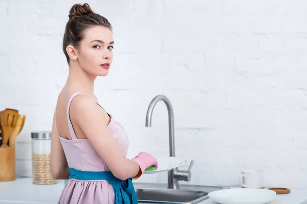 Atractiva mujer joven en guantes de goma rosa plato de lavado con esponja en la cocina - foto de stock