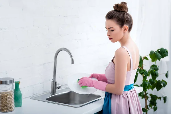 Mujer joven pensativa en guantes de goma rosa plato de lavado con esponja en la cocina - foto de stock