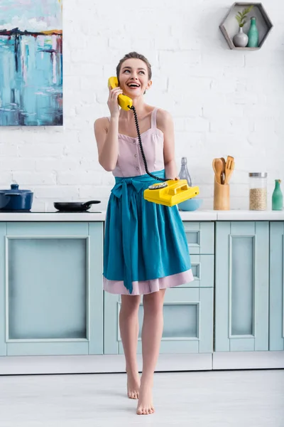 Счастливая босиком молодая женщина разговаривает по ретро желтый телефон и смеется на кухне — стоковое фото