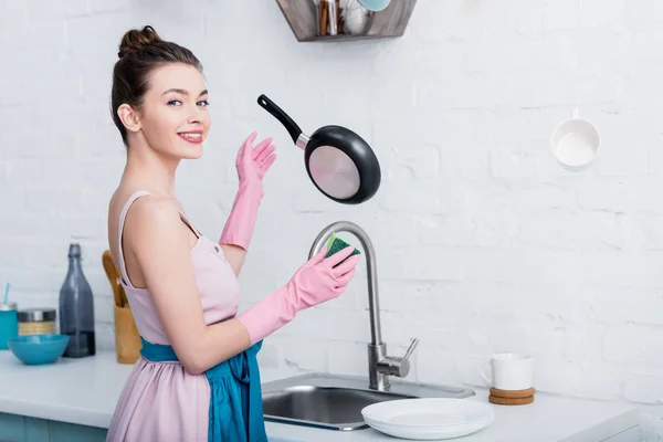 Sonriente joven feliz con guantes de goma mirando a la cámara mientras cocina utensilios levitando en el aire - foto de stock