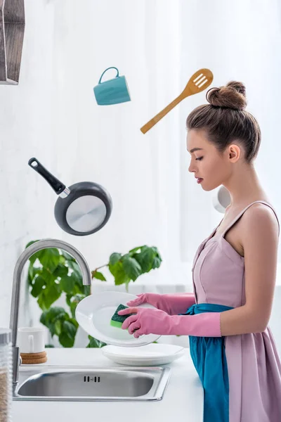 Jovem mulher em luvas de borracha lavar pratos enquanto cozinha utensílios levitando no ar — Fotografia de Stock