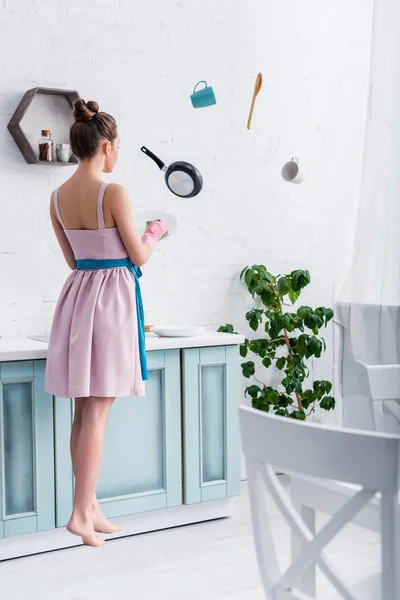 Mujer joven en guantes de goma levitando en el aire con utensilios de cocina en la cocina mientras lava la placa - foto de stock