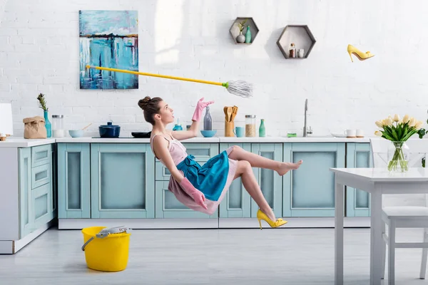 Chica elegante en delantal levitando con fregona y zapato de tacón amarillo durante la limpieza de la casa en la cocina - foto de stock