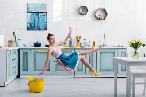 Shocked girl in apron levitating with mop and yellow heeled shoe during house cleaning in kitchen — Stock Photo