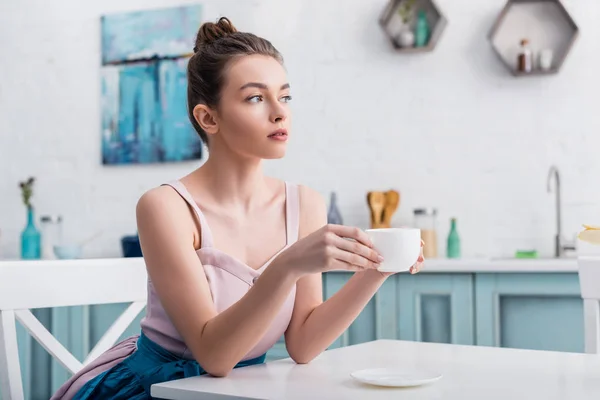 Rêveuse belle jeune femme assise à table, buvant du café et regardant ailleurs — Photo de stock