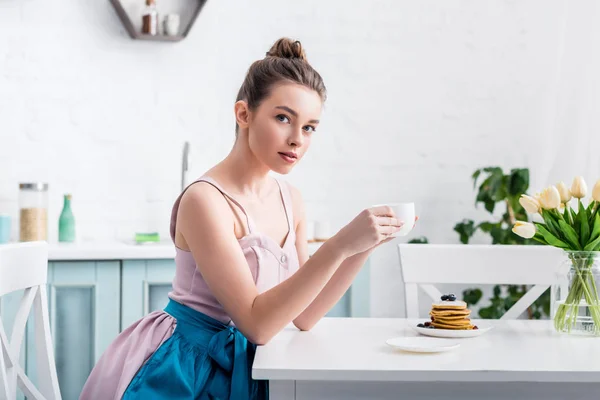 Мечтательная красивая молодая женщина сидит за столом и пьет кофе на кухне — стоковое фото