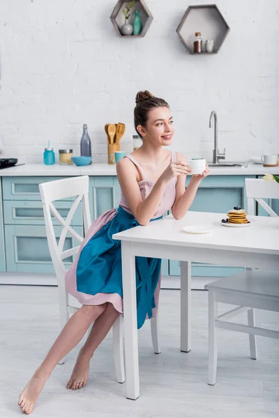 Улыбаясь босиком красивая молодая женщина сидит за столом и завтракает — стоковое фото