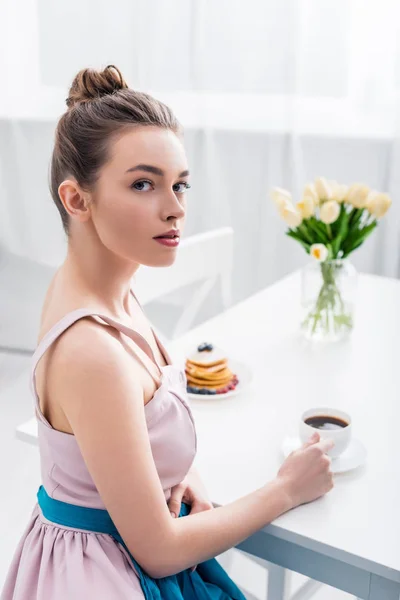 Привлекательная элегантная молодая женщина сидит за столом с тюльпанами и блинами и пьет кофе — стоковое фото
