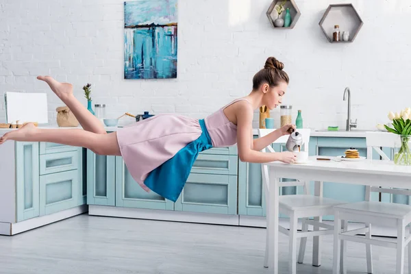 Hermosa mujer joven levitando en el aire y vertiendo té en la taza de la tetera en la cocina - foto de stock