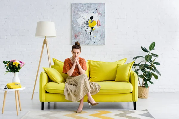 Chère élégante jeune femme assise avec les jambes croisées et les mains serrées sur le canapé jaune dans le salon — Photo de stock