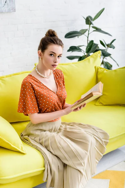 Atraente jovem mulher ler livro enquanto sentado no sofá amarelo brilhante — Fotografia de Stock