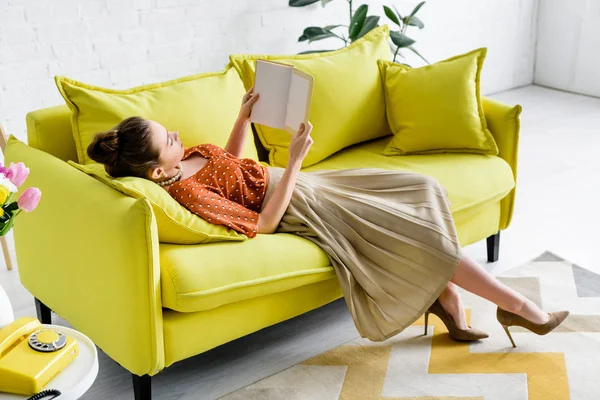 Elegant young woman resting on yellow sofa while reading book — Stock Photo