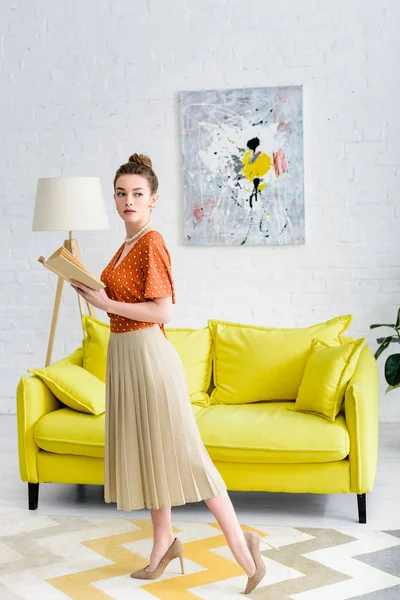 Attraktive, elegante junge Frau hält Buch in der Hand und schaut weg, während sie im Wohnzimmer steht — Stockfoto