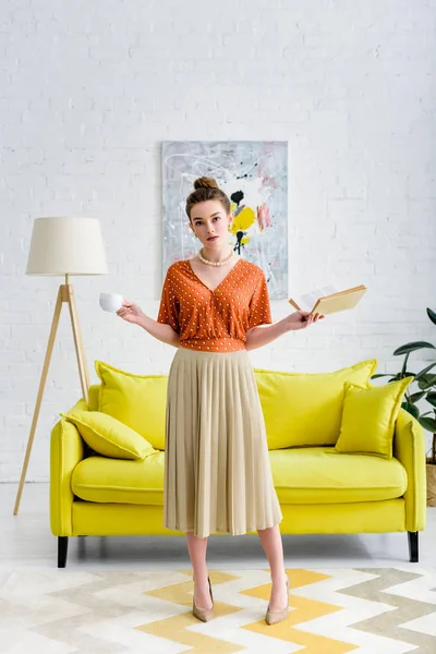 Attractive elegant young woman holding book and coffee cup in living room — Stock Photo