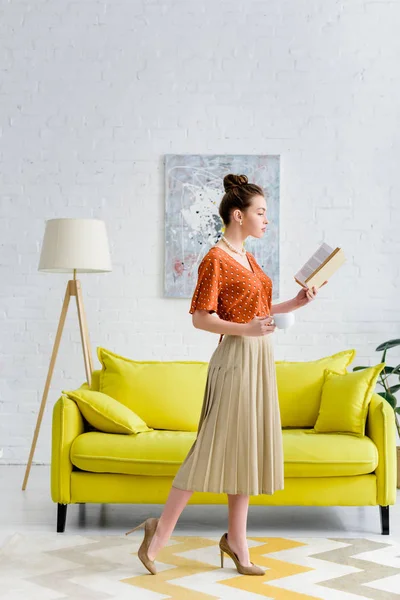 Attractive elegant young woman reading book and holding coffee cup in living room — Stock Photo