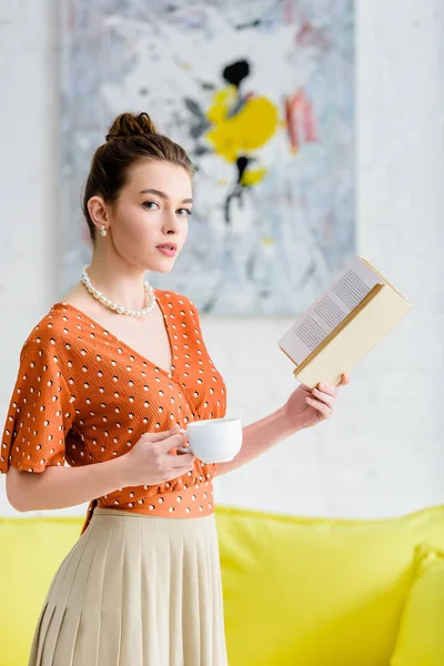 Елегантна молода жінка в перловому намисті тримає книгу і чашку, дивлячись на камеру — стокове фото