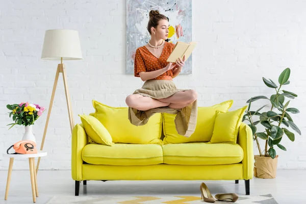 Элегантная молодая женщина в позе лотоса левитирует в воздухе во время чтения книги в гостиной — стоковое фото