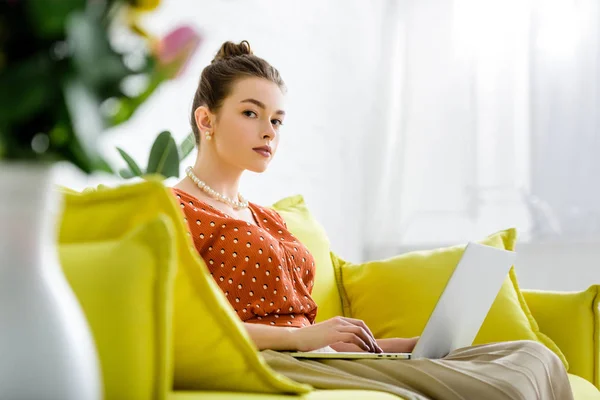 Selective focus of fashionable young woman in pearl necklace sitting on yellow sofa and using laptop — Stock Photo