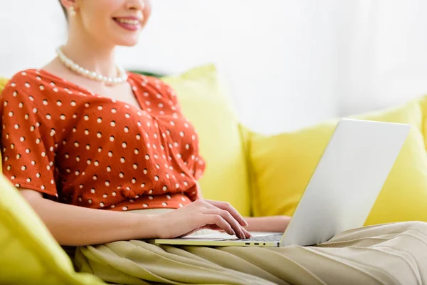 Vista recortada de la joven sonriente en collar de perlas sentado en el sofá amarillo y el uso de la computadora portátil - foto de stock