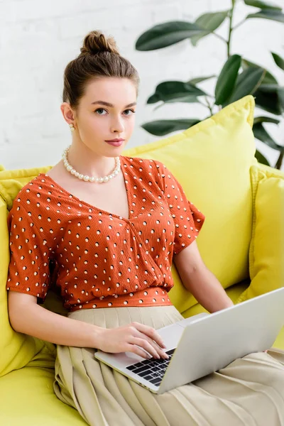 Selbstbewusste modische junge Frau in Perlenkette sitzt auf gelbem Sofa und benutzt Laptop — Stockfoto
