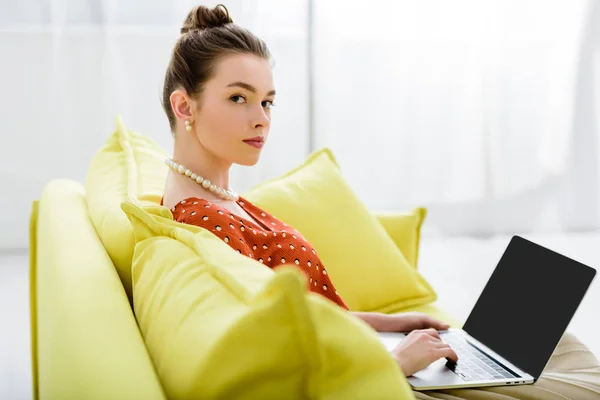 Fiduciosa elegante giovane donna in collana di perle seduta sul divano giallo e utilizzando il computer portatile con schermo bianco — Foto stock