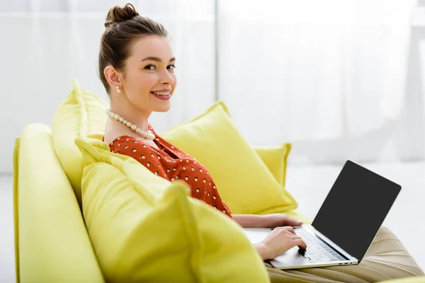 Усміхнена елегантна молода жінка в перловому намисті сидить на жовтому дивані і використовує ноутбук з порожнім екраном — стокове фото