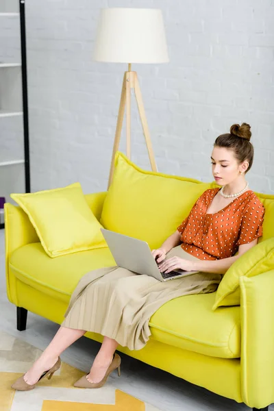 Elegante junge Frau sitzt auf gelbem Sofa und benutzt Laptop im Wohnzimmer — Stockfoto