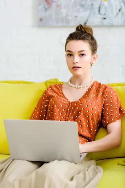 Jovem mulher elegante confiante sentado no sofá amarelo, usando laptop na sala de estar e olhando para a câmera — Fotografia de Stock