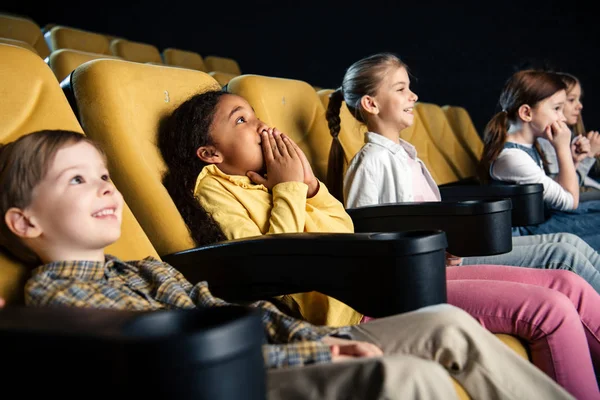 Sorrindo amigos multiculturais sentados no cinema e assistindo filme juntos — Fotografia de Stock