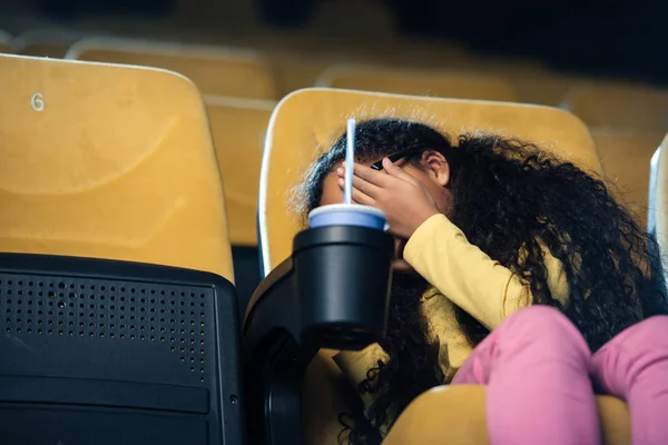 Enfant afro-américain effrayé tenant la main sur le visage alors qu'il était assis sur le siège du cinéma avec une tasse en papier dans le porte-gobelet — Photo de stock