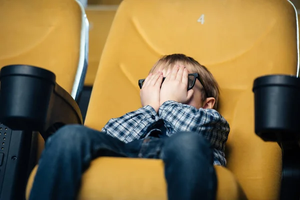 Garçon effrayé tenant la main sur le visage tout en étant assis au cinéma et en regardant le film — Photo de stock